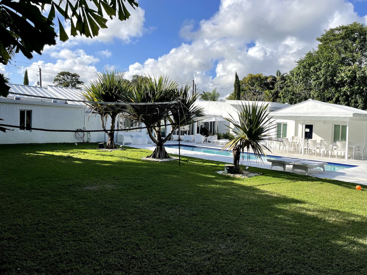 A spacious backyard features a large heated pool surrounded by well-maintained grass and palm trees. White lounge chairs line the pool area, and the house is visible in the background, showcasing modern architecture and ample windows.
