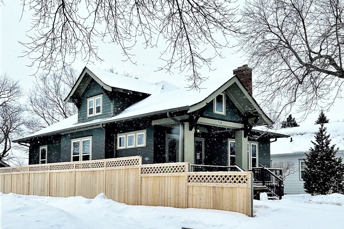 A historic Craftsman-style home, covered in snow, stands amidst a serene winter landscape. The exterior features intricate woodwork and large overhangs. A snow-dusted wooden fence encloses a portion of the yard, while trees are bare against the cloudy sky.