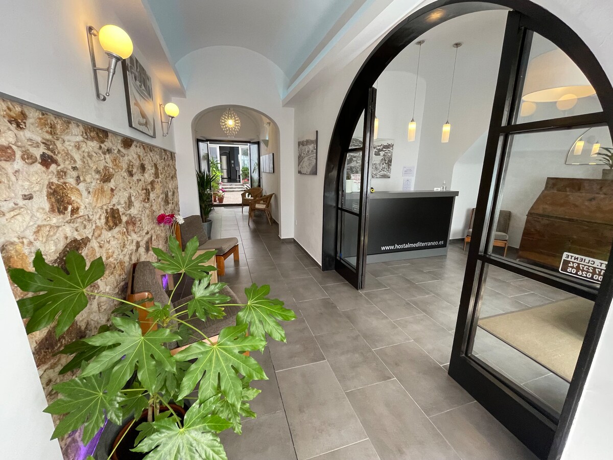 The welcoming lobby area features smooth stone flooring and arched doorways leading to separate spaces. Natural light enters through large windows, revealing a reception desk and comfortable seating. Greenery is present, adding a touch of nature to the environment.