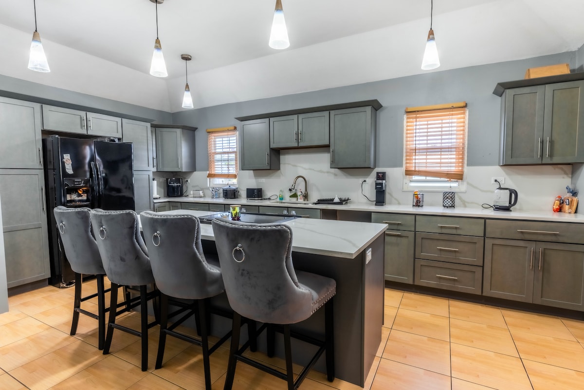 An upgraded kitchen is showcased with modern cabinetry and an island. Four high-backed chairs are positioned at the island, complemented by stainless steel appliances and natural light from a window with wooden blinds.