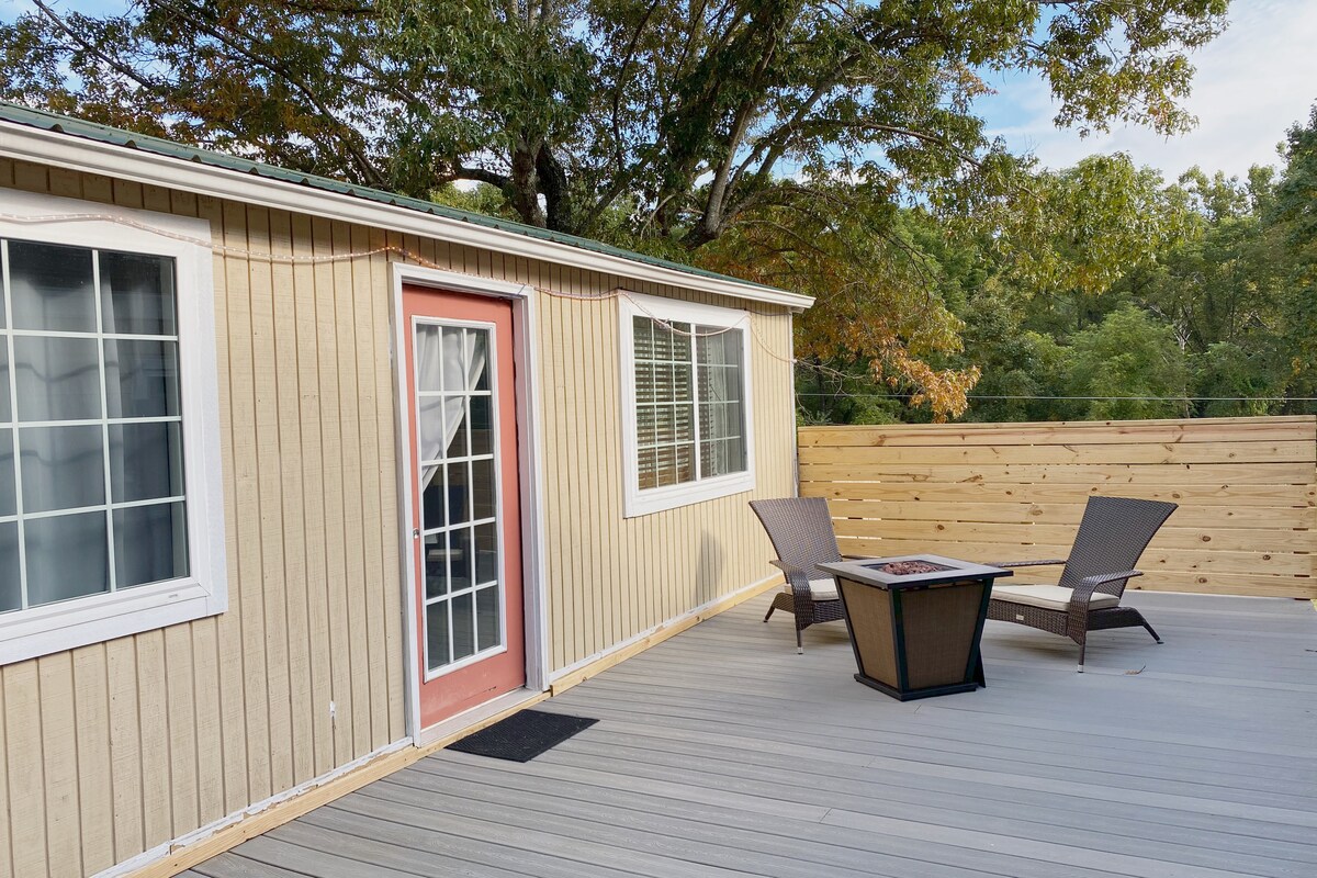 A private outdoor deck area features a small fire pit surrounded by two chairs. The guesthouse's entrance is highlighted by a pink door, and ample natural light streams through the large windows. A wooden privacy fence encircles the space, providing a secluded atmosphere.