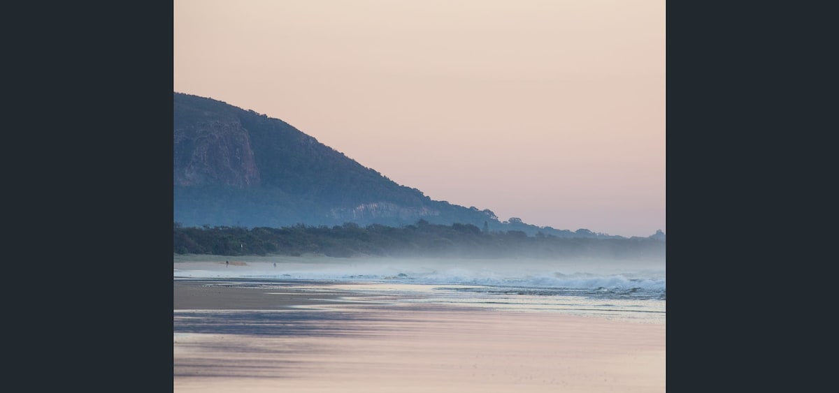 A tranquil coastal scene is presented, featuring gentle waves lapping at a sandy shore. Soft pastel colors of dawn are reflected in the water, while a distant mountain range creates a serene backdrop amidst lightening fog.