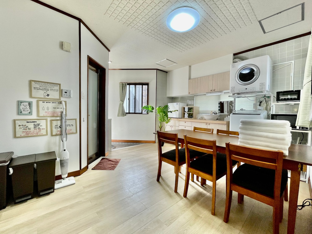 A clean and well-furnished living space features a dining table with several wooden chairs and ample natural light from a nearby window. A washing machine is positioned in the kitchen area, complemented by essential appliances for guest convenience.