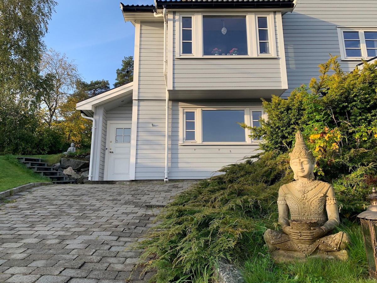 The exterior of a modern house is showcased, featuring a light-colored facade and multiple large windows. A serene statue is positioned near the entrance, surrounded by lush greenery and a stone pathway leading to the door.