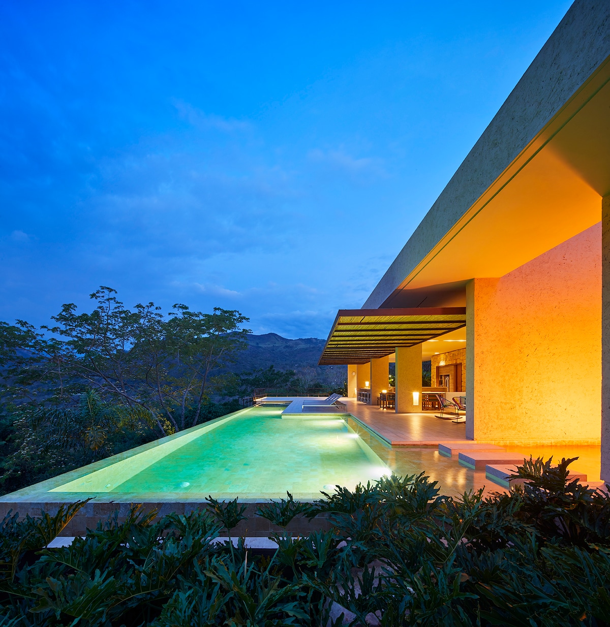 An illuminated infinity pool stretches alongside the modern structure, reflecting the evening sky. The surrounding greenery enhances the serene environment, while the open terrace invites relaxation. Soft lighting from the house highlights the architectural features, creating a tranquil ambiance.