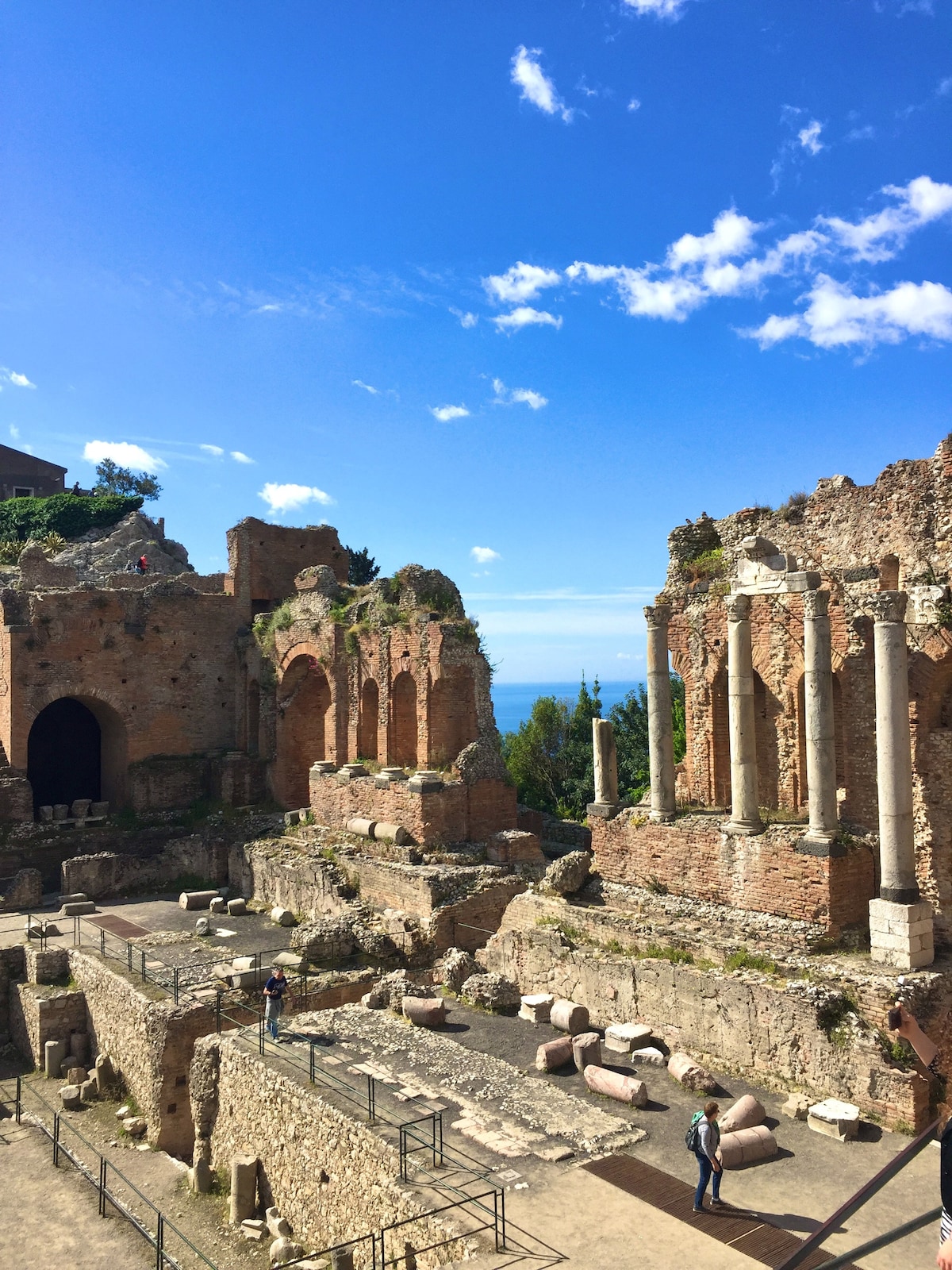 Ancient ruins are depicted with remnants of stone columns and arches, surrounded by lush greenery. The background features a clear blue sky and a glimpse of the sea, while visitors can be seen exploring the historic site.