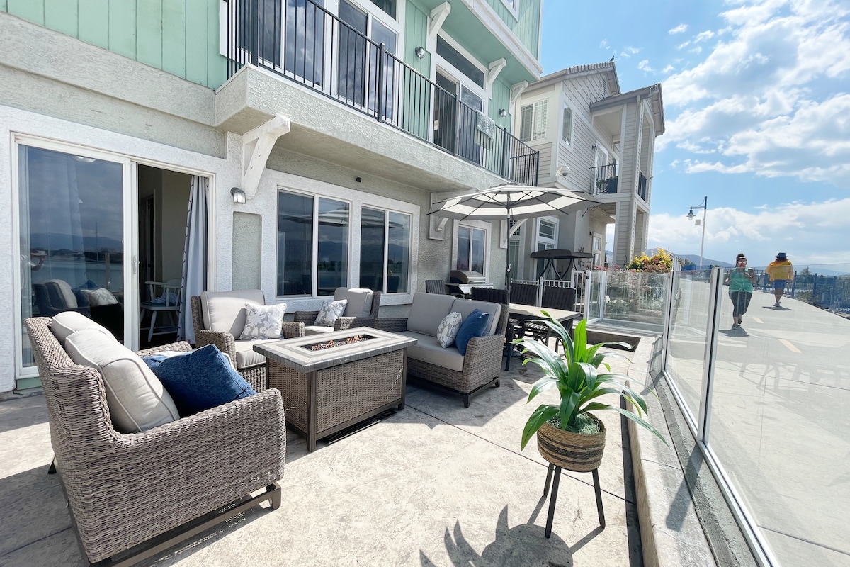 An outdoor patio features comfortable seating arrangements, including a sofa and armchairs, surrounding a central fire table. An umbrella provides shade, with scenic views of the lake and mountains visible in the background. Glass barriers enhance the open feel of the space.