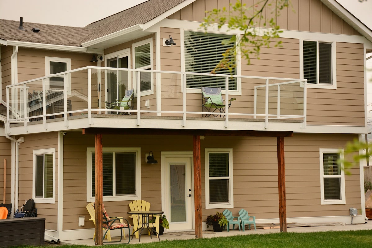 A two-story house with modern farmhouse decor is depicted, featuring a glass-railed balcony on the upper floor. The lower level has a patio area with colorful outdoor seating and large windows, allowing for ample natural light. A well-maintained lawn is visible in front.