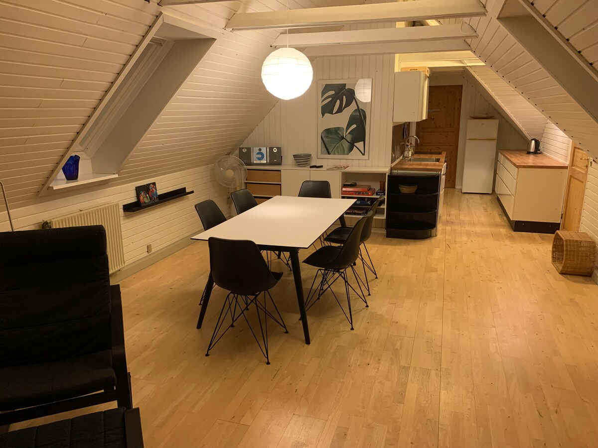 A spacious high-ceilinged room is illuminated by a pendant light. A dining table surrounded by six chairs occupies the center, while a small kitchen area is visible in the background. Light wood flooring complements the modern design, and a wall art piece is displayed nearby.