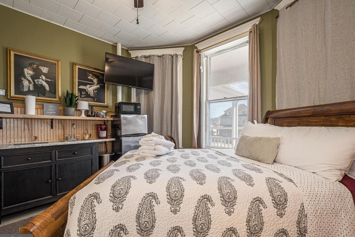 The studio features a queen bed with a light-colored blanket and decorative pillows. A television is mounted on the wall, and a small fridge is visible nearby. Two large windows allow natural light to fill the space, complemented by artworks hanging above a wooden storage unit.