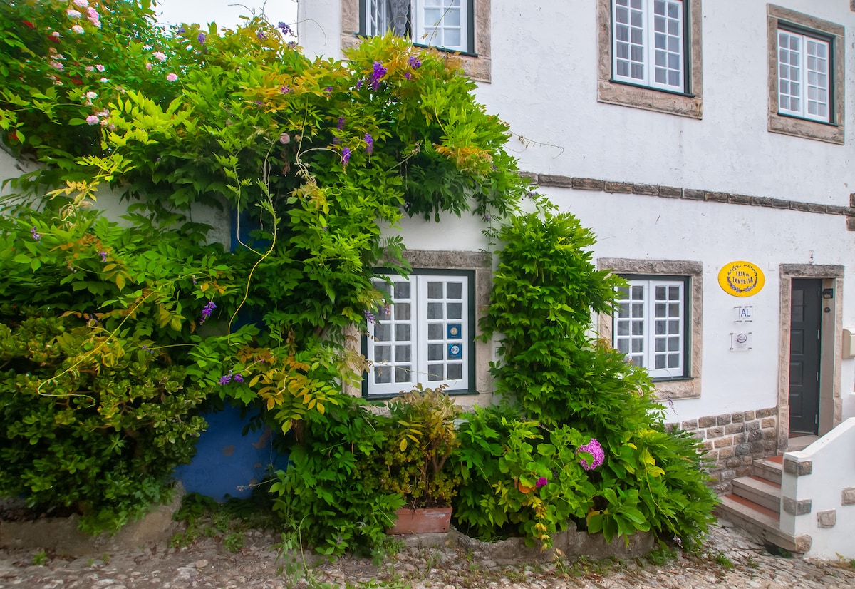The exterior of a traditional house is surrounded by lush greenery and colorful flowering plants. Multiple windows are framed by vibrant foliage, creating a welcoming entrance. The stone pathway leads to the door, enhancing the quaint charm of the home within Óbidos.