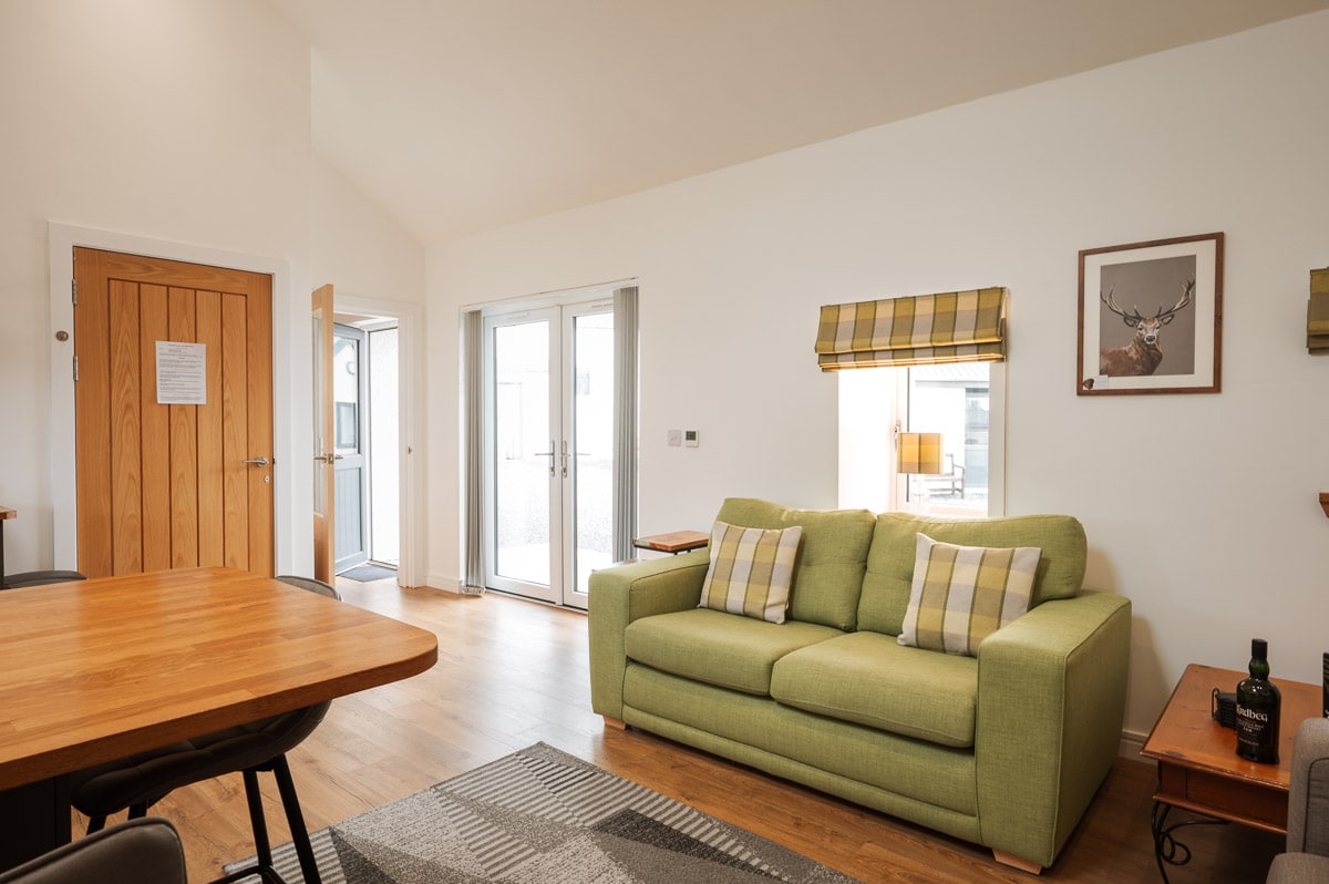 An open plan living area features a green sofa with patterned cushions, complemented by a wooden dining table. Natural light floods in through patio doors, which lead to the outdoor seating area. A framed deer print decorates the wall, enhancing the cozy atmosphere.