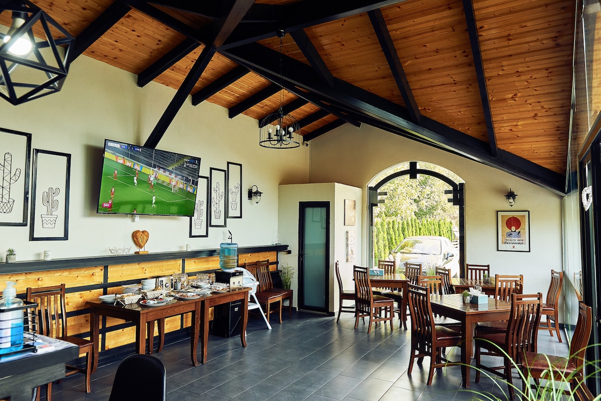 An airy dining area features wooden beams overhead and large windows allowing natural light. Several wooden tables and chairs are arranged for guests, while a television is mounted on the wall. Decorative frames enhance the space's character, creating an inviting atmosphere for meals.