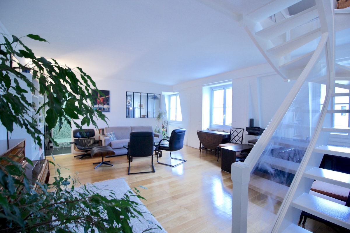 A bright living area features light wood flooring and large windows allowing natural light. Stylish furniture, including a modern lounge chair and a minimalist sofa, complements the space. A spiral staircase leads to an upper level, enhancing the open layout of the room.
