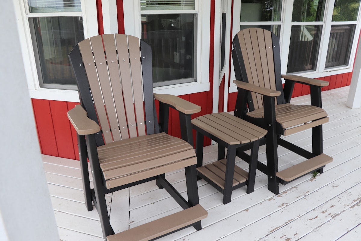 Two Adirondack chairs are positioned on a covered porch, accompanied by a small side table. The chairs feature a mix of light brown and black colors, while the porch has a white wooden floor and red walls, providing a comfortable spot to relax.