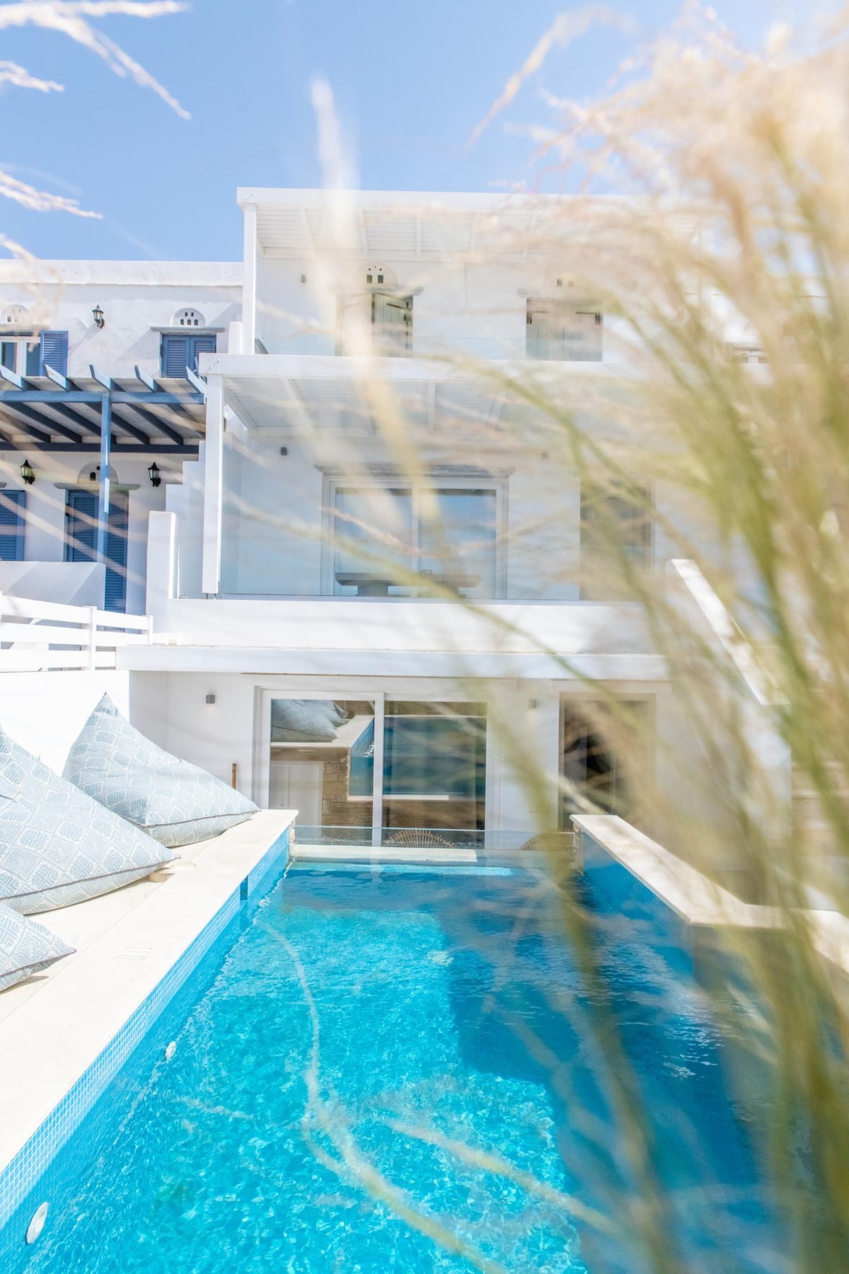 A serene view of a modern multi-level building by a swimming pool, framed by tall grass. The pool's clear blue water reflects the sunlight, while seating areas with cushions are positioned around the pool, providing a space for relaxation.
