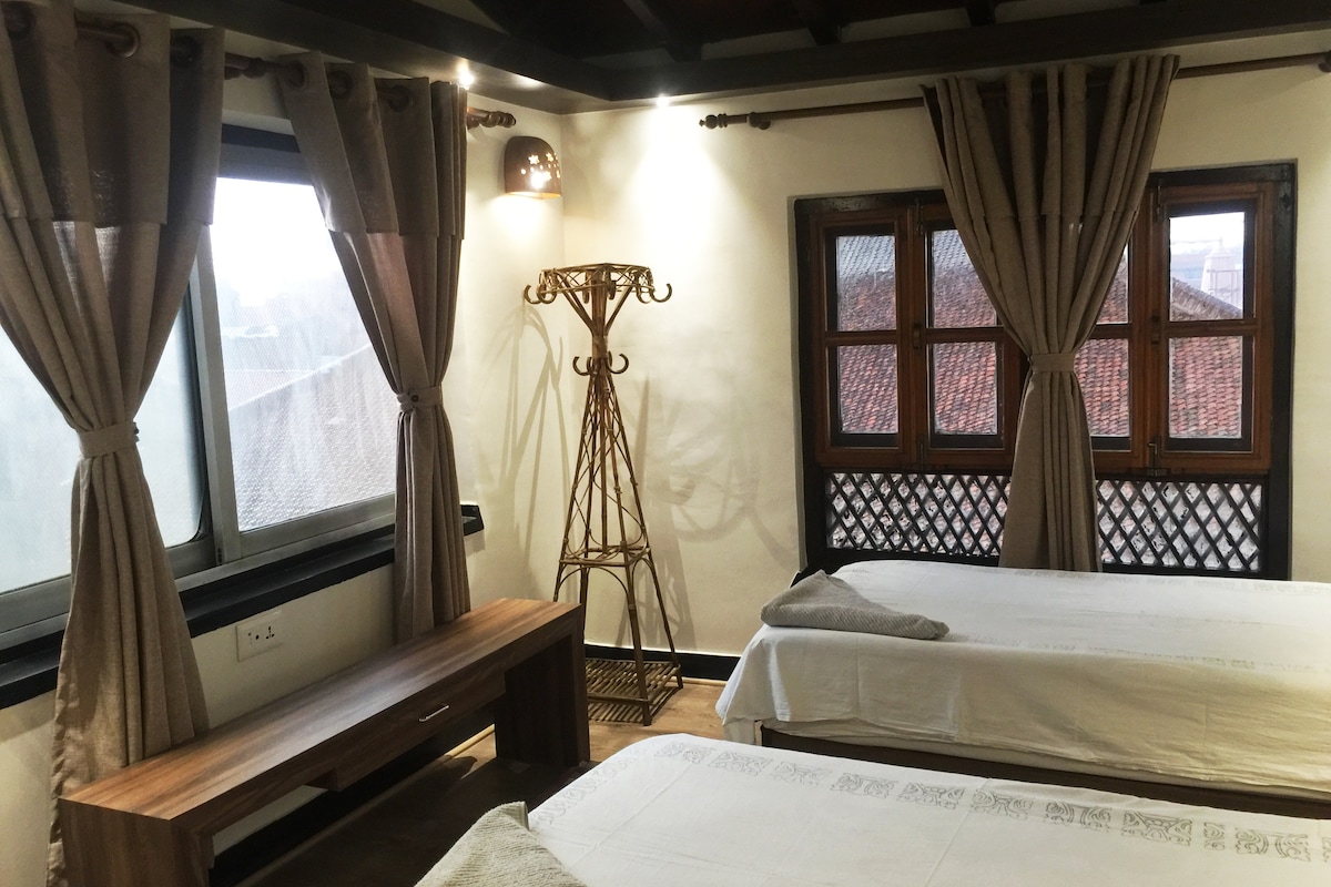 A spacious room is presented with two beds dressed in white linens. Natural light streams through large windows adorned with elegant curtains. A wooden bench is placed against one wall, while a decorative stand adds a stylish touch to the corner.