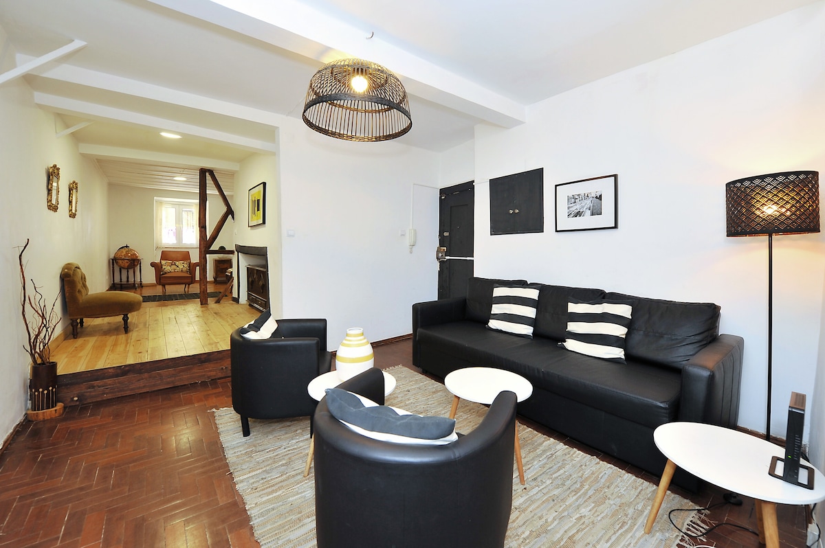 A modern living area features a black leather sofa with striped cushions, paired with two round chairs. Light-colored side tables complement the space. A decorative lamp stands next to the seating area, while wood flooring adds warmth. A separate room is visible at the back.
