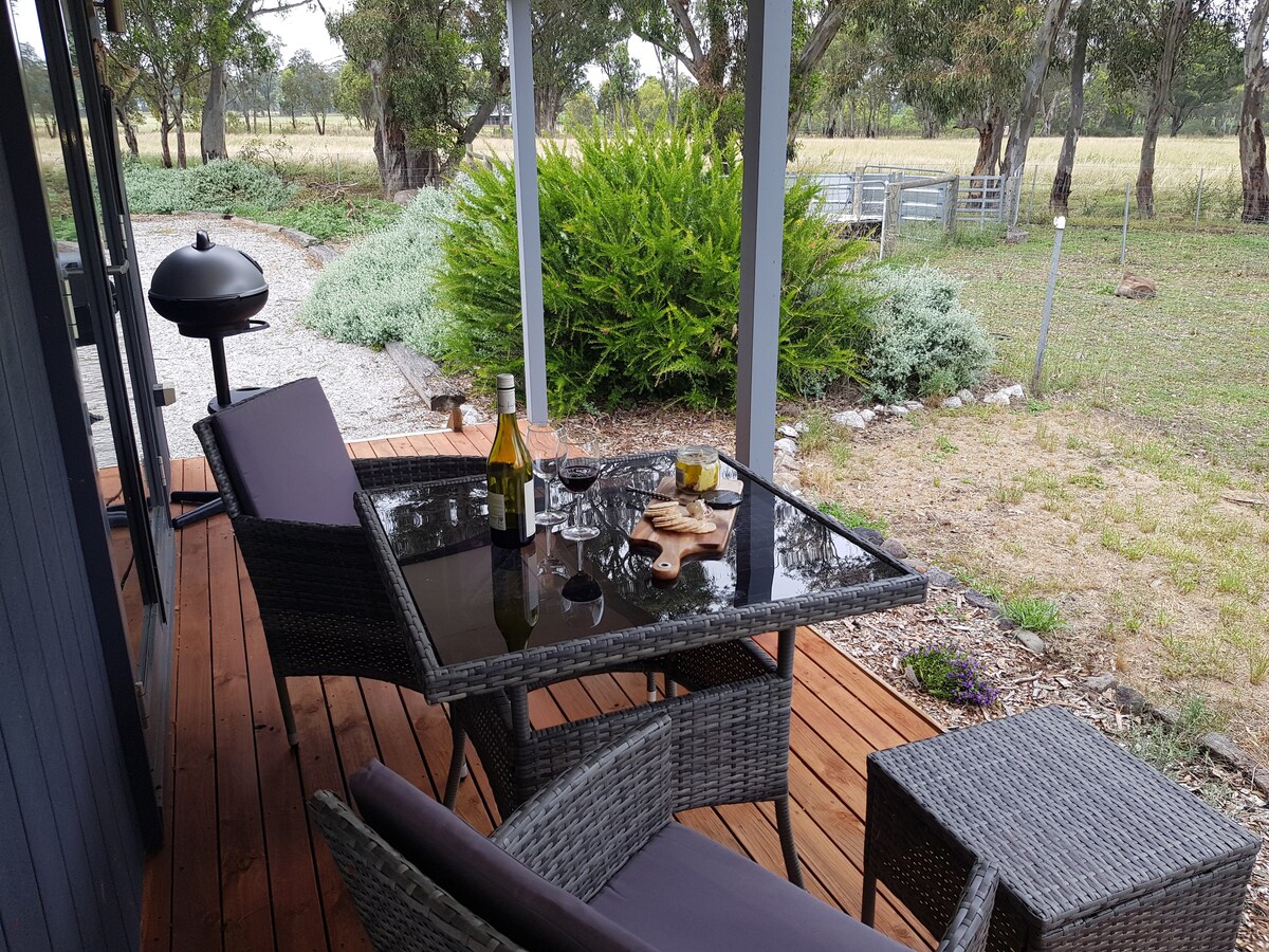 An outdoor dining area features a glass tabletop with a wine bottle, glasses, and a plate of snacks. Two comfortable chairs and a BBQ are positioned on a wooden deck, surrounded by greenery and a view of the open paddock beyond.