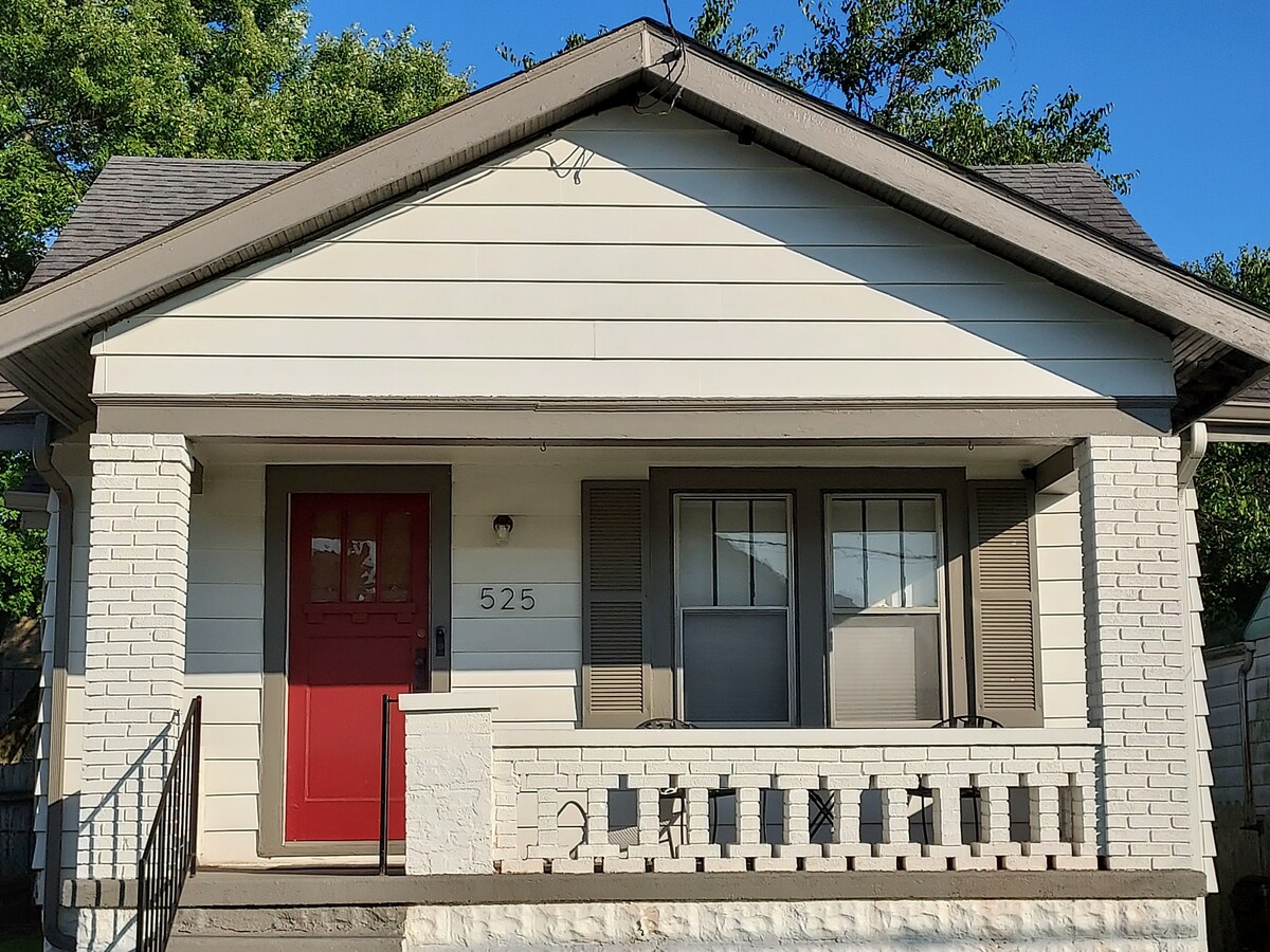 The exterior of a single-story home is presented, featuring a welcoming red door that stands out against white siding. Two front windows with shutters are visible, and a small porch with a decorative railing is framed by greenery from nearby trees.