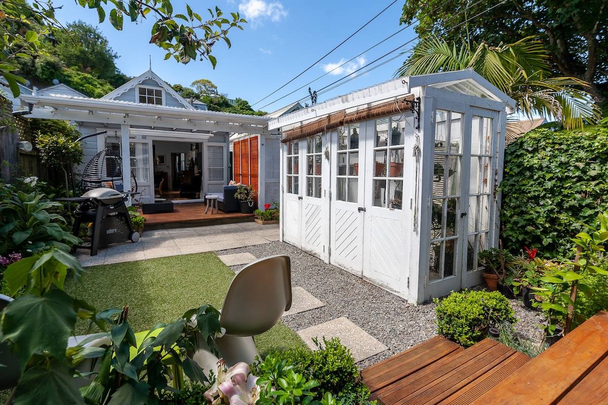 A charming outdoor space is displayed, featuring a mix of greenery and landscaped areas. A white garden shed with glass doors complements the cottage in the background. The patio includes a decorative gravel path and a small patch of grass, enhancing the inviting atmosphere.