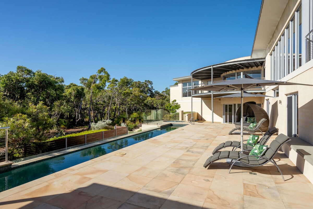 An expansive outdoor patio features a lap pool lined by modern loungers. Umbrellas provide shade beside the pool, while lush greenery surrounds the area, offering a tranquil retreat. The residence blends elegantly with the natural landscape, showcasing a seamless indoor-outdoor connection.