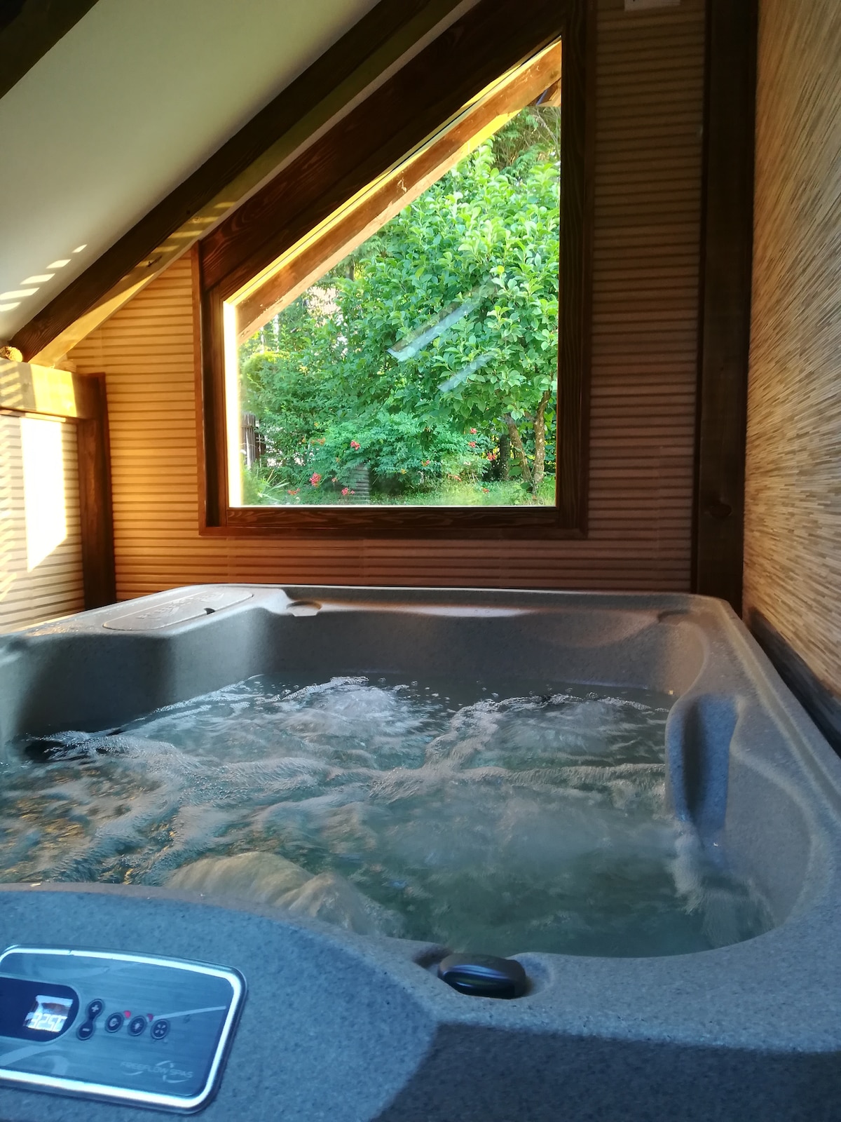 A spacious hot tub is positioned beside a large window, inviting natural light and views of the lush greenery outside. The soothing water is gently bubbling, creating a tranquil atmosphere. The warm tones of the wooden interior enhance the relaxing environment.