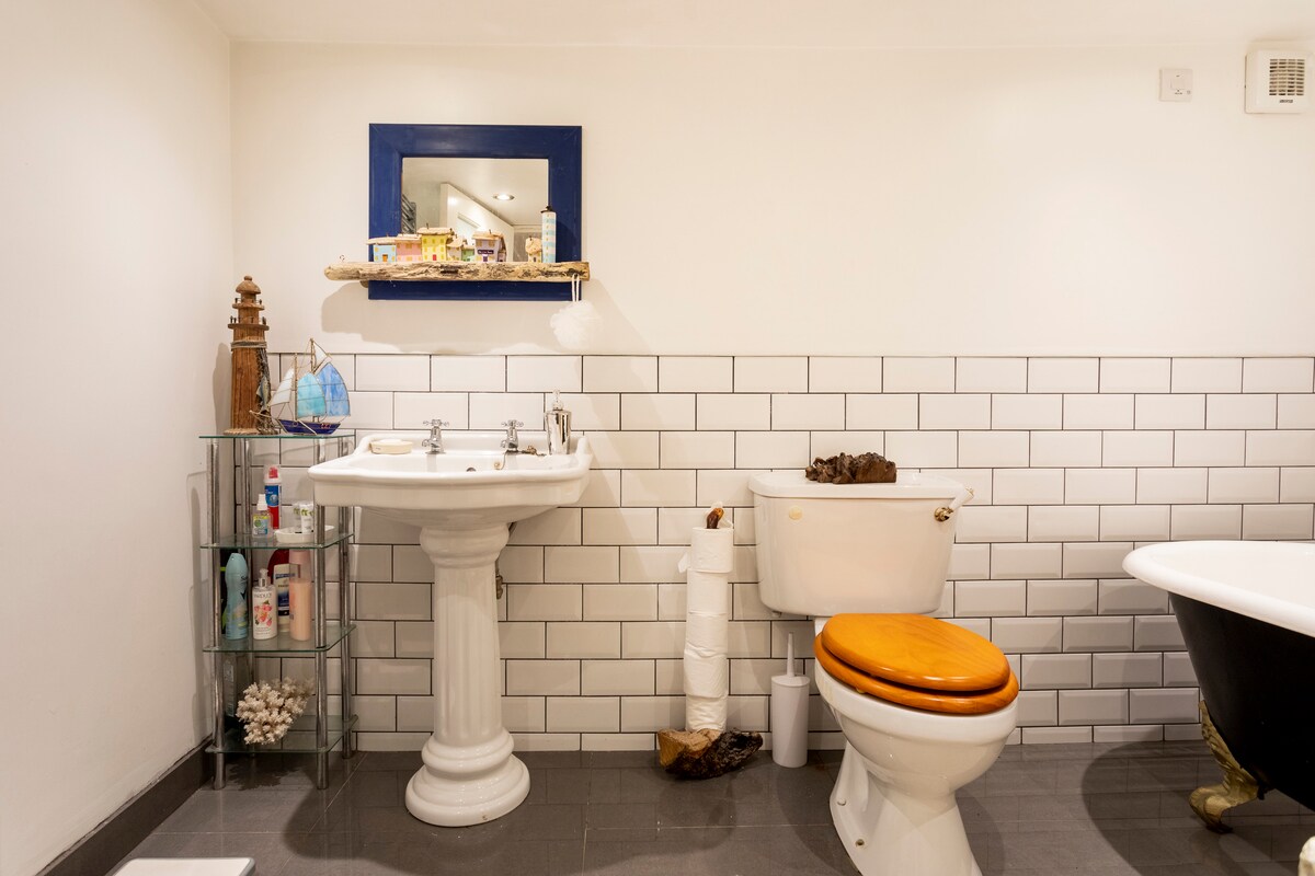 The bathroom features a classic pedestal sink and a wooden toilet seat, accompanied by a freestanding bath with a vintage design. A glass shelf holds various toiletries, while decorative elements like shells are visible. The tiled wall creates a clean and modern aesthetic.