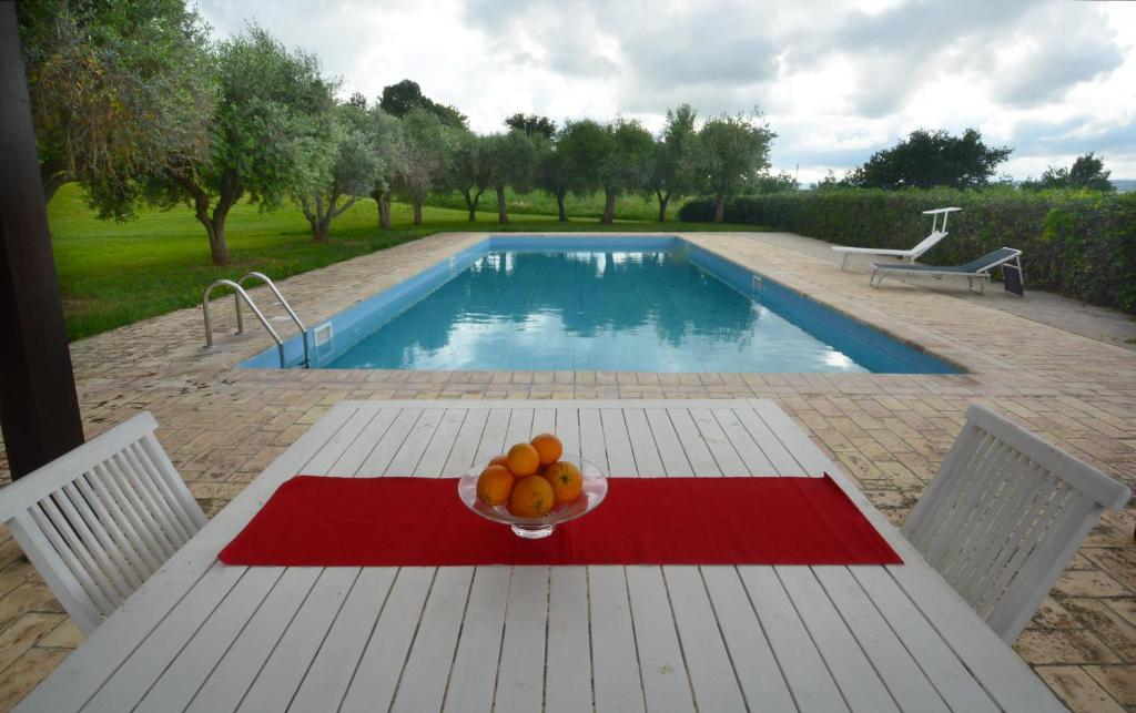 An outdoor dining space is presented, featuring a white table adorned with a vibrant red runner and a bowl of oranges. A serene swimming pool glistens in the background, surrounded by green landscaping and trees, creating a peaceful retreat.