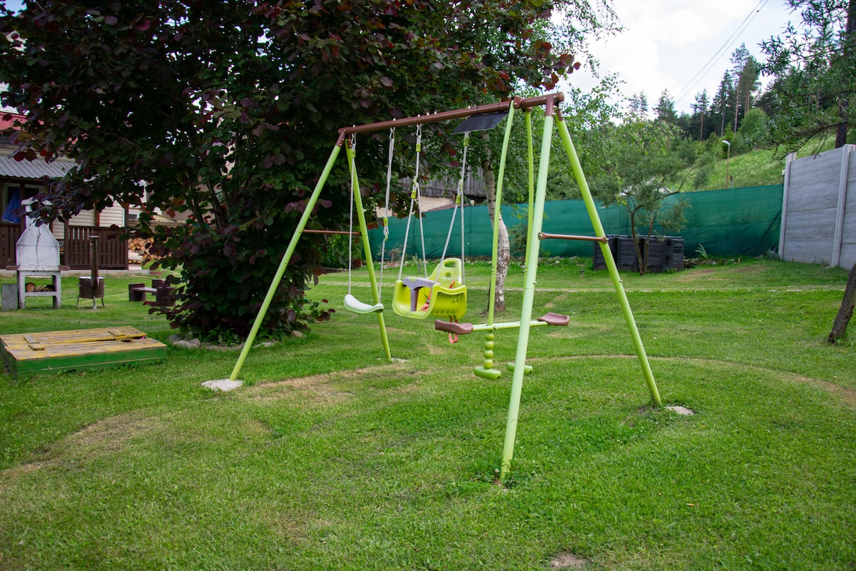 A playground area features a green swing set set upon a grassy lawn. The swing set includes child-sized swings and a baby swing, providing a safe space for children to play. Surrounding greenery enhances the natural setting.
