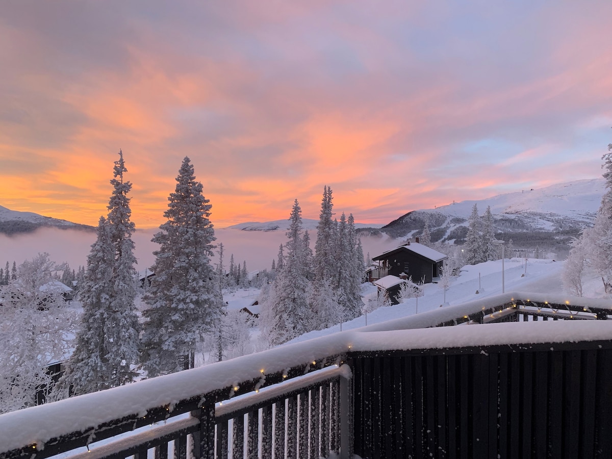 A winter landscape is displayed, with a balcony railing in the foreground adorned with soft lights. Snow-covered trees surround the view, while a vibrant sunset casts shades of pink and orange across the sky, illuminating the lower layers of snow and creating a serene atmosphere.