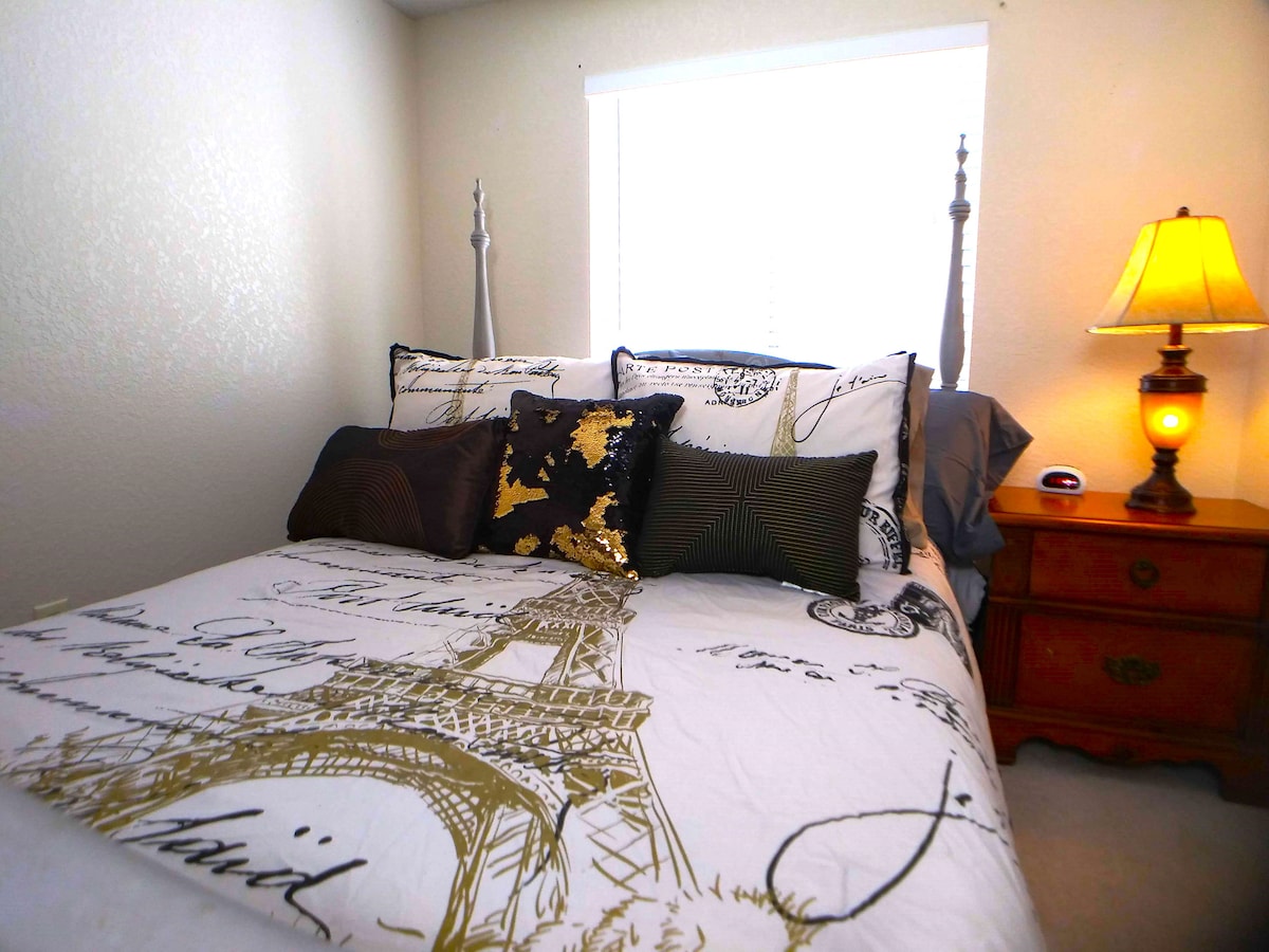 A cozy sleeping area features a spacious bed adorned with an artistic patterned duvet cover and multiple decorative pillows. A bedside lamp casts a soft glow, while a window allows natural light to filter in, complemented by light-colored walls.