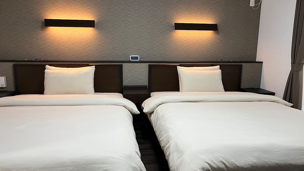 Two spacious double beds are positioned side by side, each adorned with cream-colored linens and plush pillows. Soft wall-mounted lights provide gentle illumination above the headboards, enhancing the restful ambiance of the room.