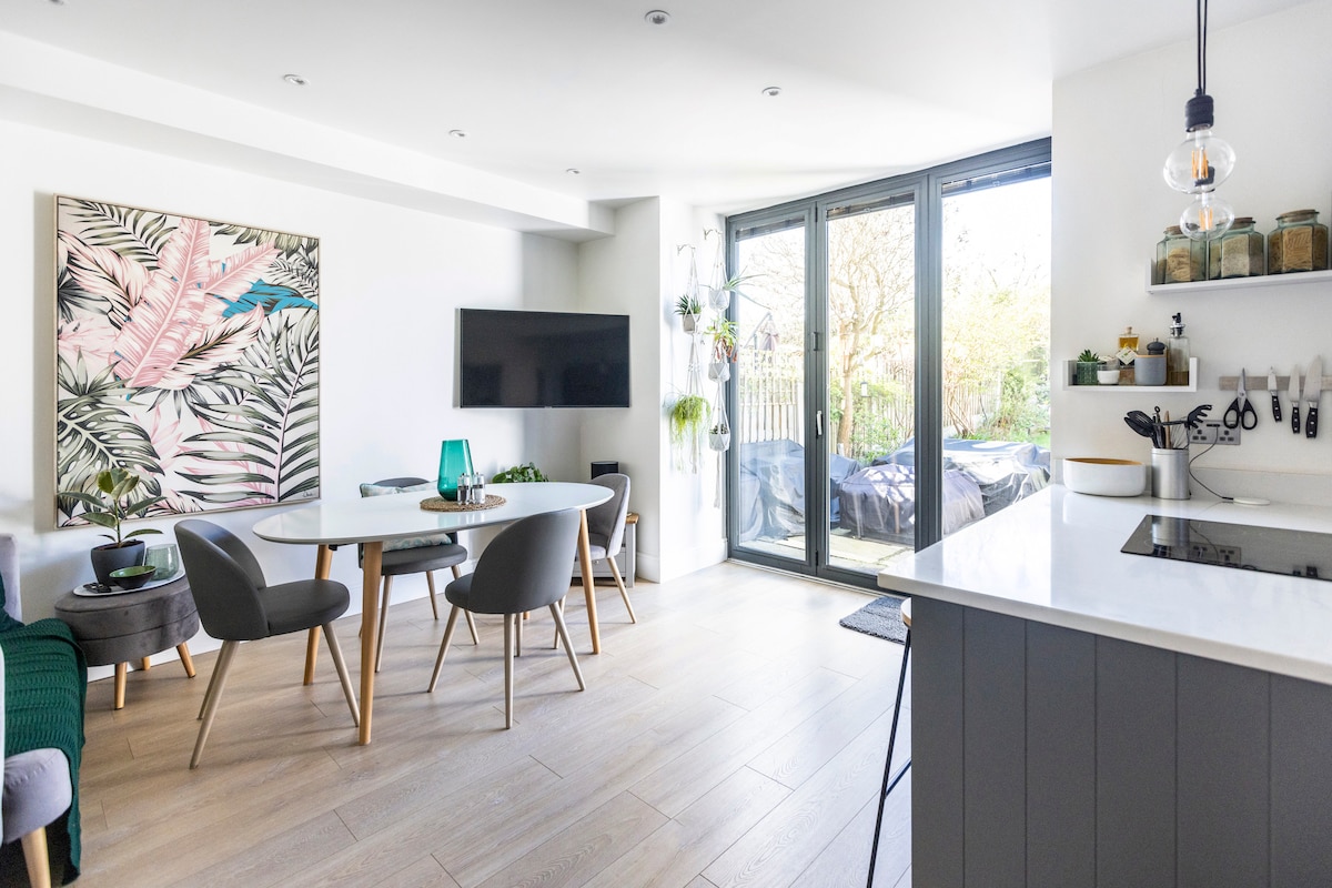 A modern dining area features a round table surrounded by four chairs and a large wall-mounted television. Floor-to-ceiling glass doors provide views of the outdoor space, while an abstract artwork adds a touch of color to the neutral interior.