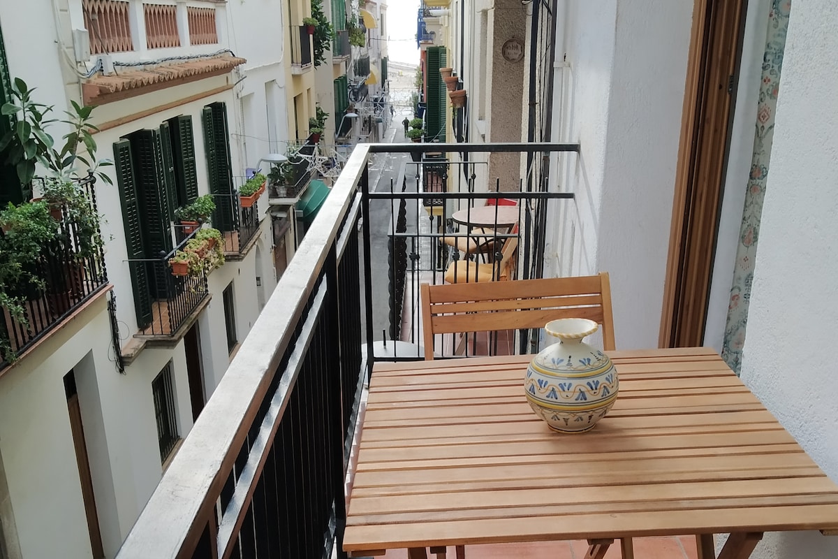 A small balcony features wooden furniture, including a table and chairs. Decorative potted plants can be seen on surrounding balconies, and the view showcases a narrow street with white-washed buildings, highlighted by green shutters and vibrant flower boxes.