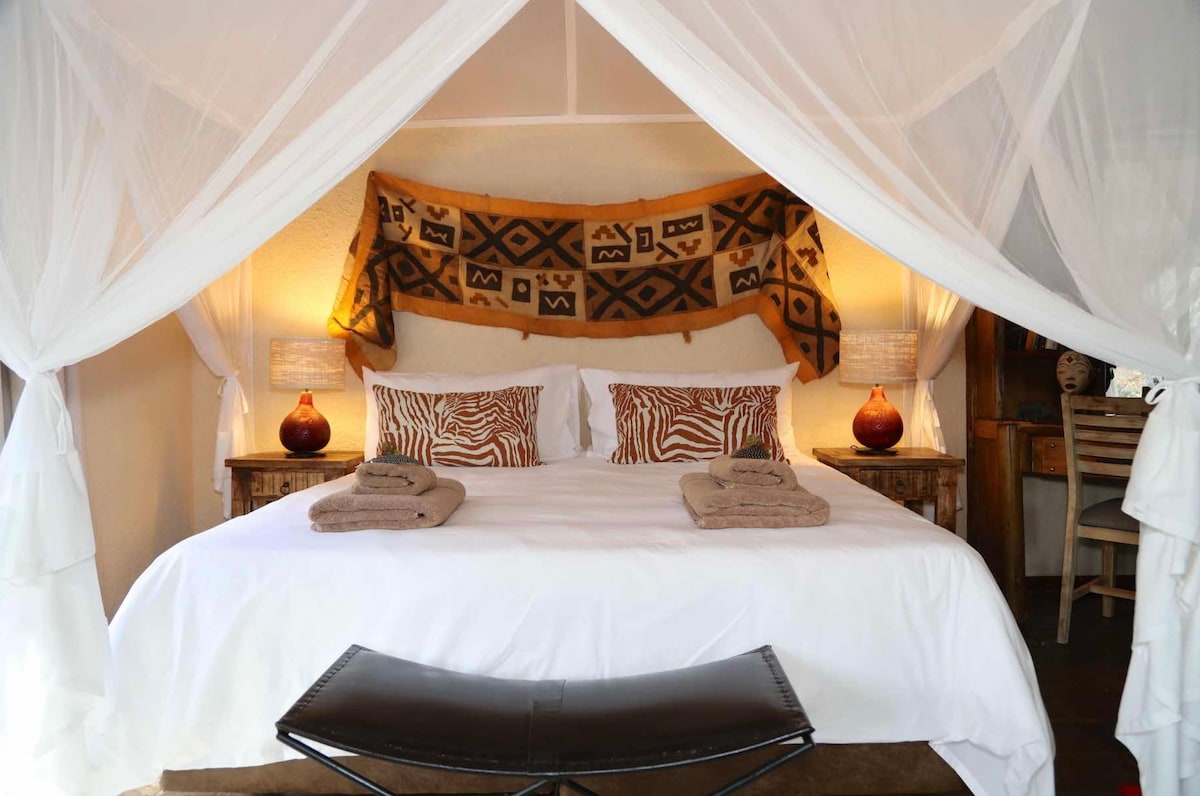A spacious sleeping area is framed by a delicate canopy, highlighting a king size bed adorned with patterned pillows. Twin lamps beside the bed create gentle lighting, while a decorative piece hangs overhead. A comfortable chair and towel sets are neatly arranged for convenience.