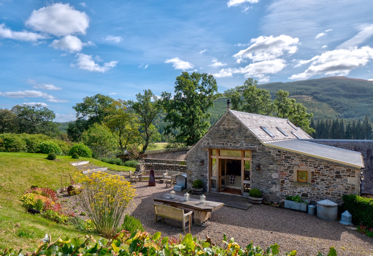 A spacious stone house is set against a backdrop of trees and hills. Outdoor seating areas are arranged around the house, including a central table, creating inviting spots for gatherings. Lush greenery and colorful flowers enhance the natural surroundings under a clear blue sky.
