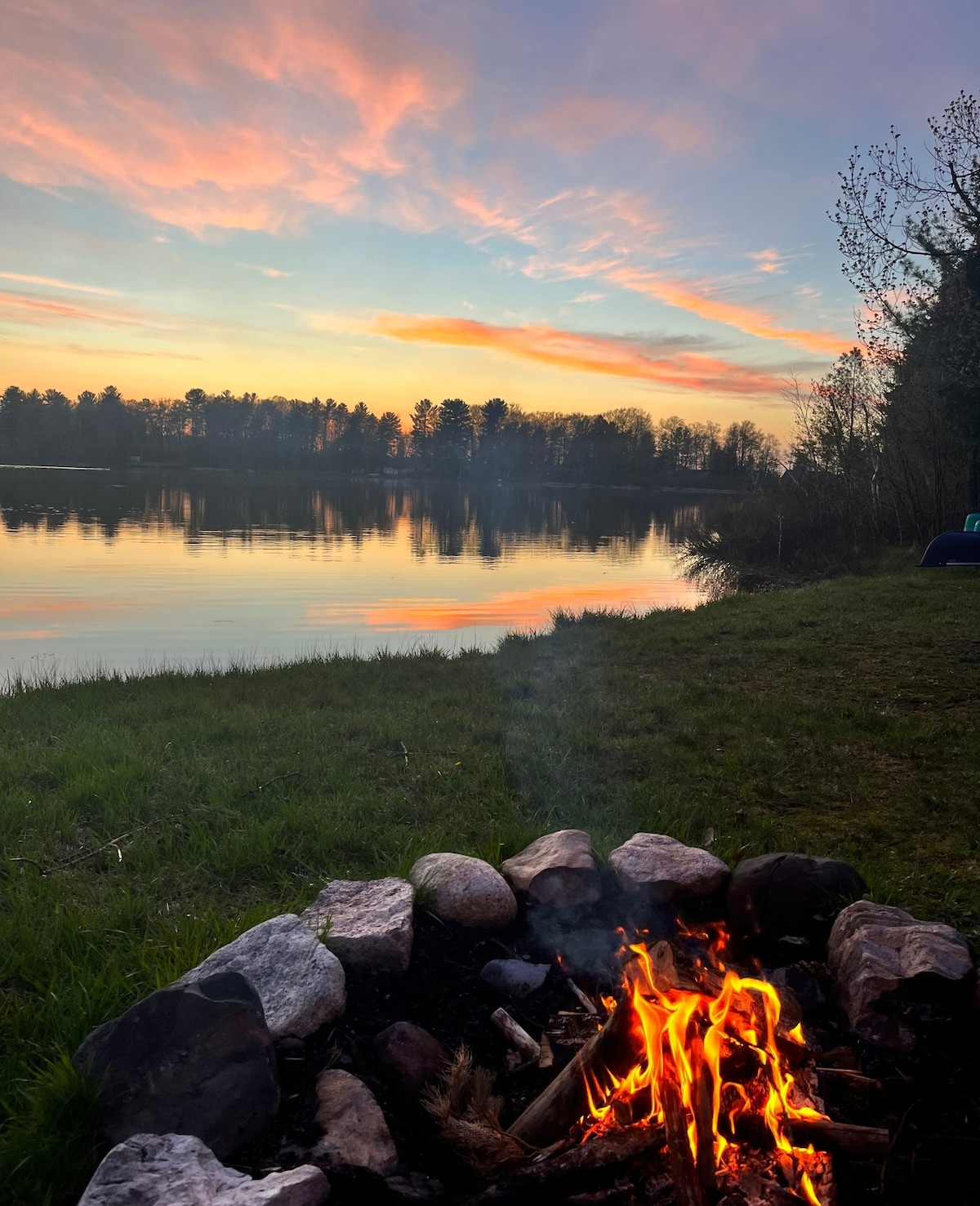 A crackling fire is enclosed within a circular stone firepit, with flames visible against the backdrop of a tranquil lake. Soft pastels from the sunset reflect on the calm water, and trees provide a serene border to the scene.