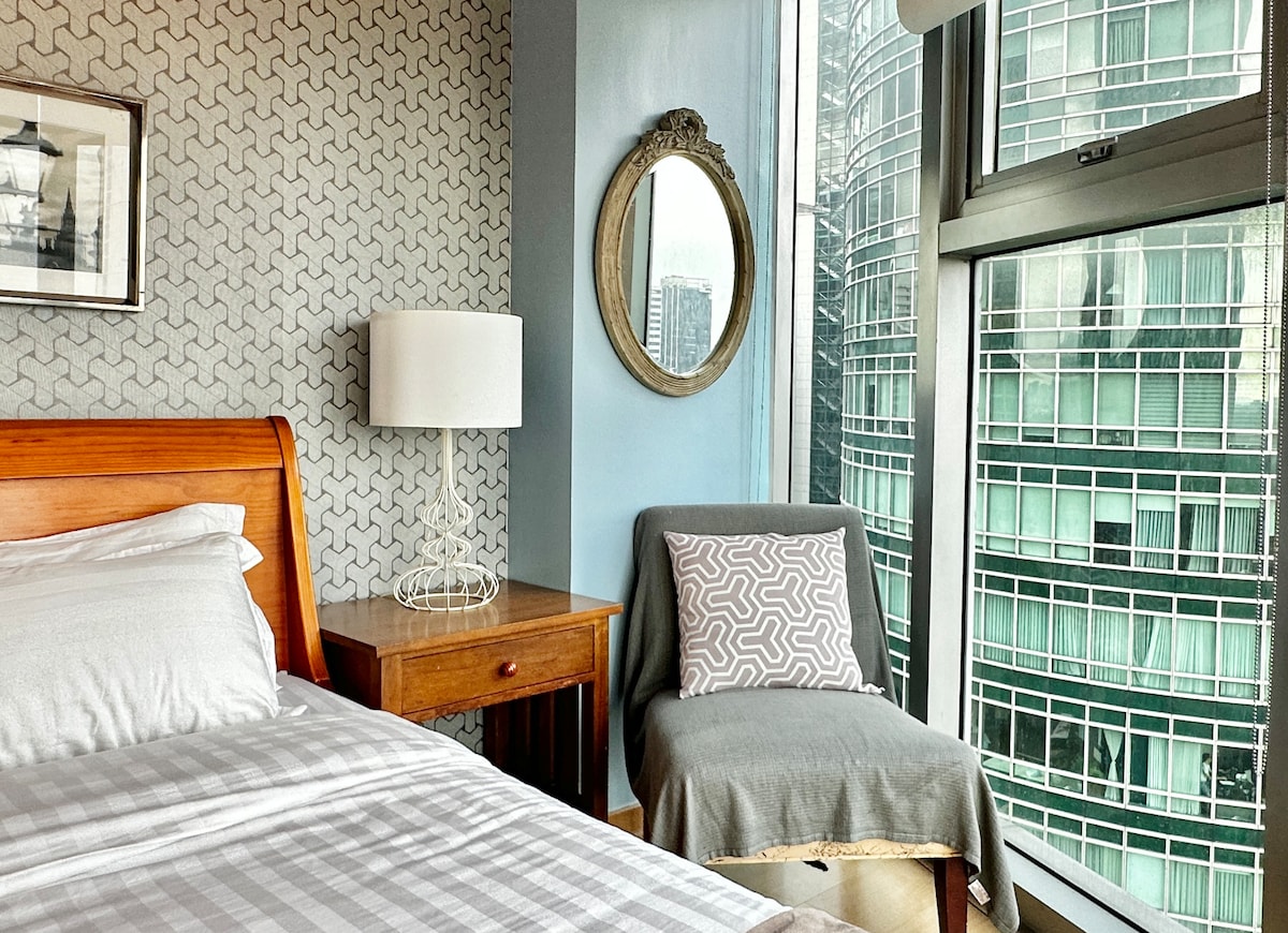 The intimate bedroom features a wooden bed with a neatly arranged white bedding set. A cozy chair is situated by the floor-to-ceiling windows, offering a view of the cityscape. A round mirror and a bedside lamp add to the room's charm.