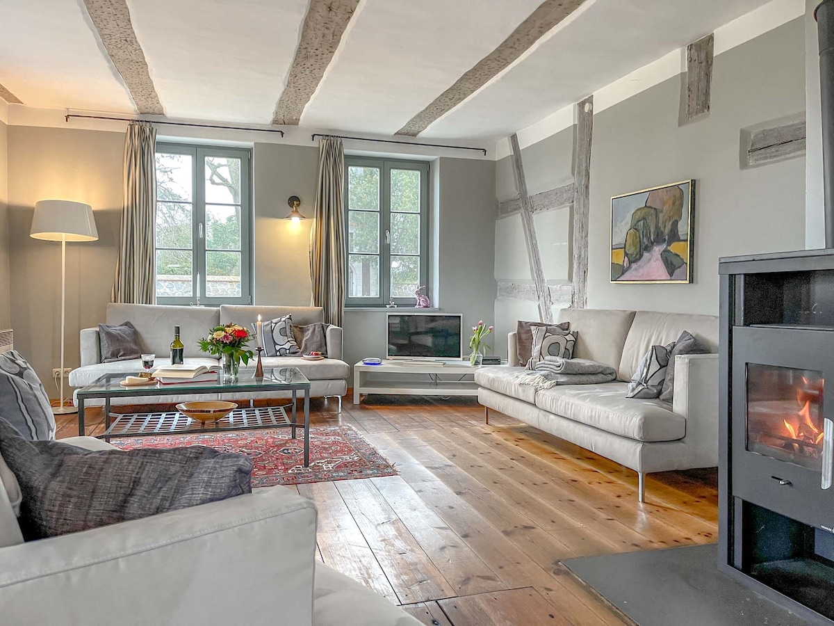 The living area features a neutral-toned decor, highlighted by a cozy seating arrangement on a wooden floor. A contemporary fireplace adds warmth, while large windows provide natural light and views of the surrounding greenery. An art piece and floral arrangement enhance the space.