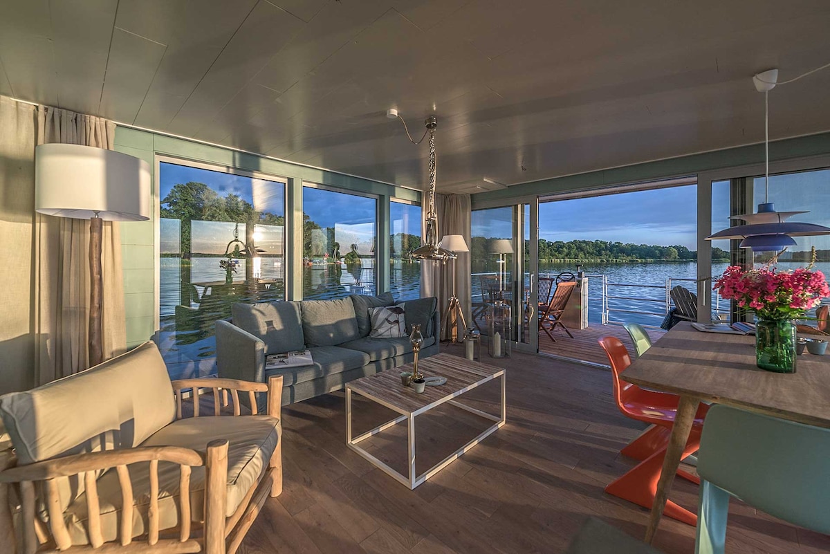 A spacious living area is featured, showcasing large windows that offer expansive views of the water. A comfortable seating arrangement with light-colored furniture is provided, along with a dining table. Natural light floods the space, creating a bright and airy ambiance.