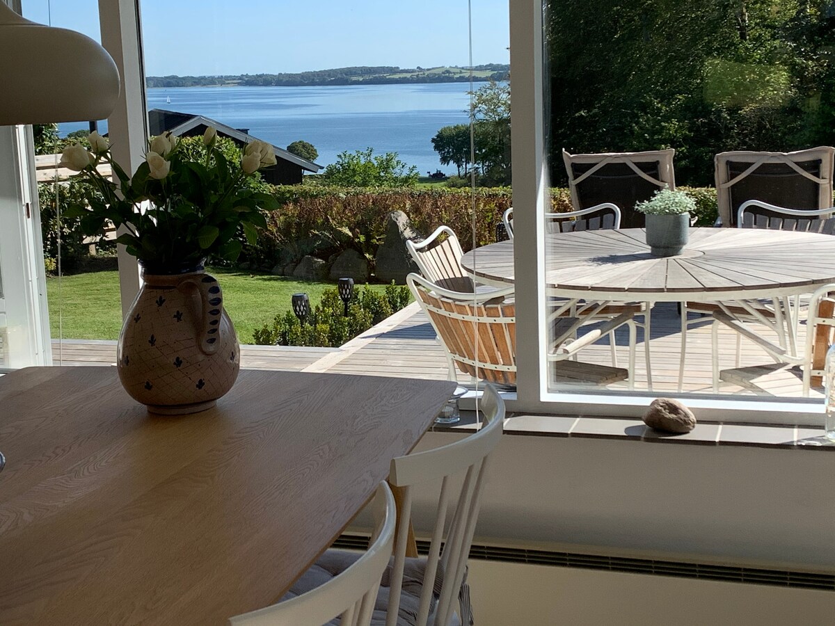 A spacious dining area features a wooden table surrounded by white chairs, with a vase of flowers as a centerpiece. A large window offers a view of a serene outdoor terrace and the water beyond, framed by greenery and bright sunlight.