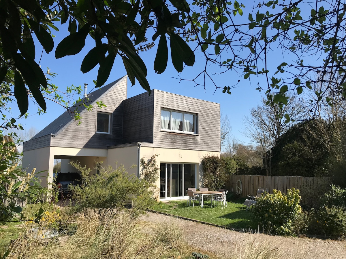 A modern house is set within a natural landscape, featuring large windows that allow light to fill the interior. A patio with outdoor dining furniture is visible, surrounded by mature shrubs and trees, creating a pleasant outdoor space. A clear blue sky completes the scene.