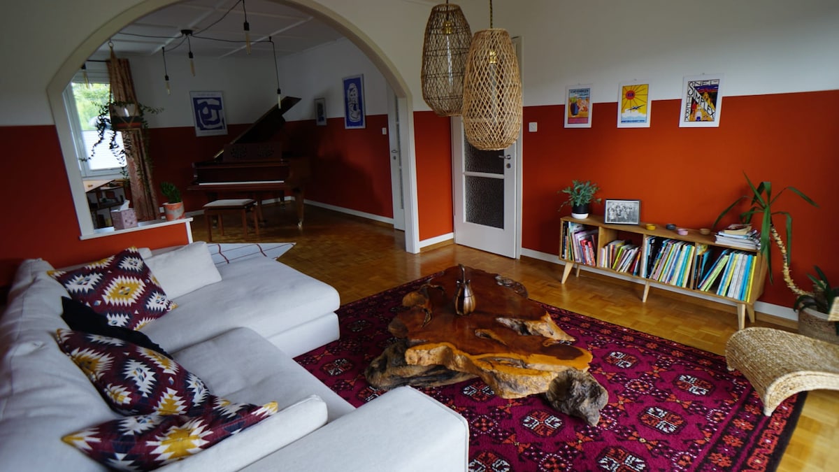 The spacious living area features an inviting white sectional sofa, a unique wooden coffee table, and a vibrant area rug. A grand piano is visible in the background, accompanied by decorative wall art and a bookshelf showcasing various titles. Natural light streams through windows, enhancing the warm ambience.