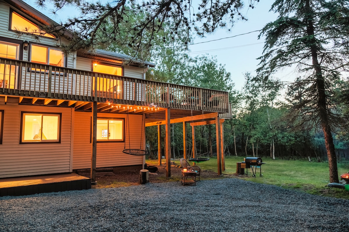 An inviting outdoor area features a spacious wraparound deck with warm lighting. The lower section includes a gravel pathway leading to seating options and a charcoal grill, surrounded by lush greenery. The house's windows glow softly as evening approaches, enhancing the tranquil atmosphere.