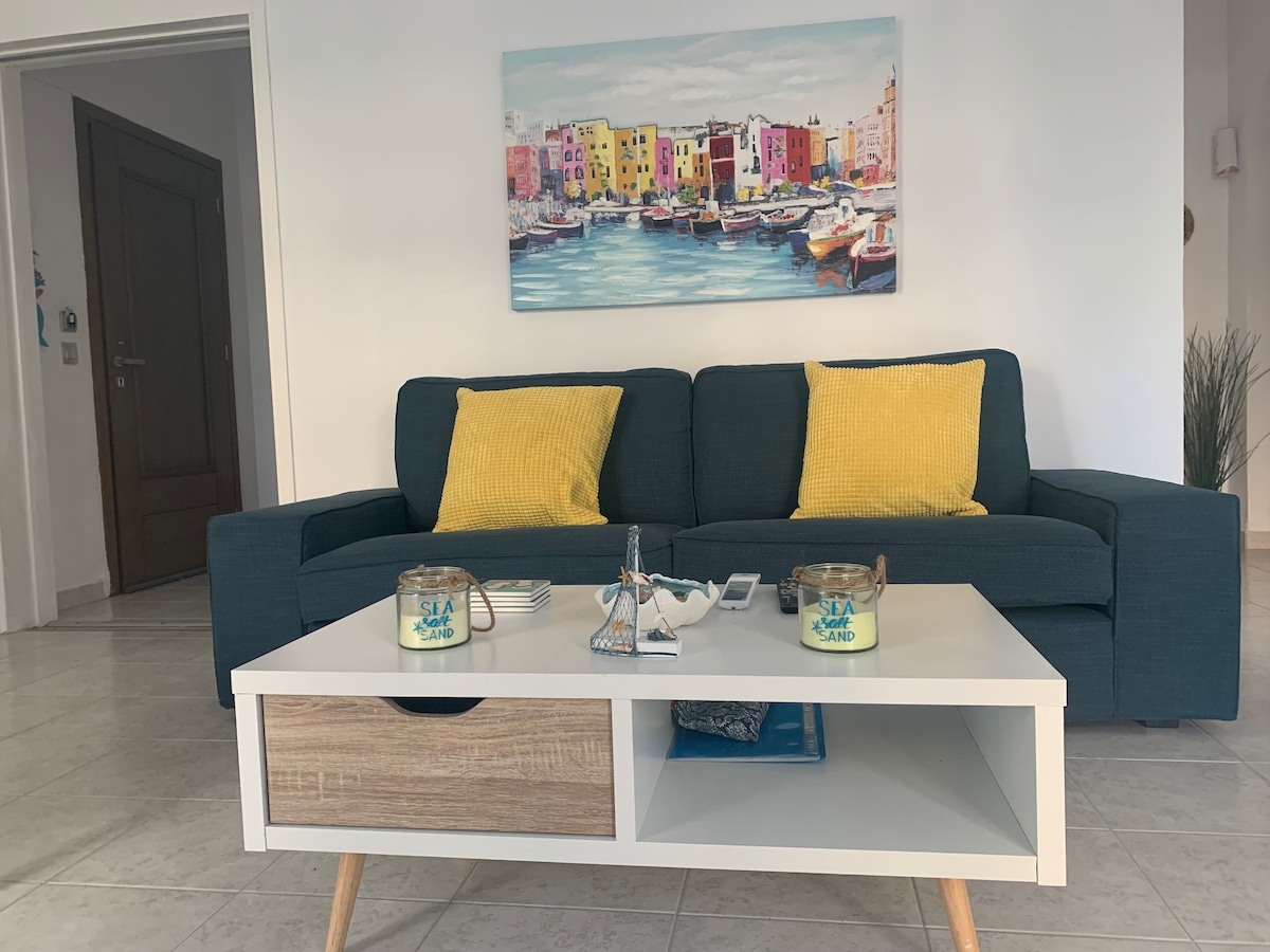 A spacious living area is depicted, featuring a comfortable dark blue sofa adorned with yellow decorative pillows. A white coffee table with a wooden accent is positioned in front, showcasing a decorative item and two small containers. A colorful artwork depicting a seaside scene hangs on the wall.