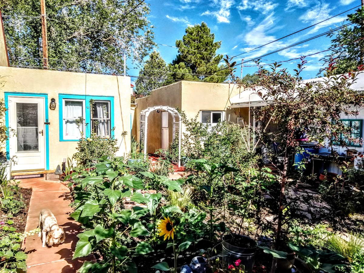 The image depicts a charming outdoor space featuring adobe-style casitas surrounded by lush greenery. A pathway of terracotta tiles leads through vibrant plants and flowers, with a bright sunflower visible in the foreground. The sky is clear with scattered clouds, enhancing the tranquil atmosphere.