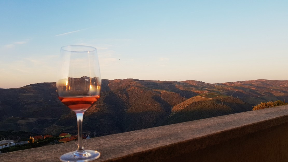 A wine glass sits elegantly on a stone railing, capturing the golden hues of the setting sun. In the background, rolling hills extend toward the horizon, dotted with vineyards. The serene sky transitions from blue to warm tones, enhancing the tranquil atmosphere.