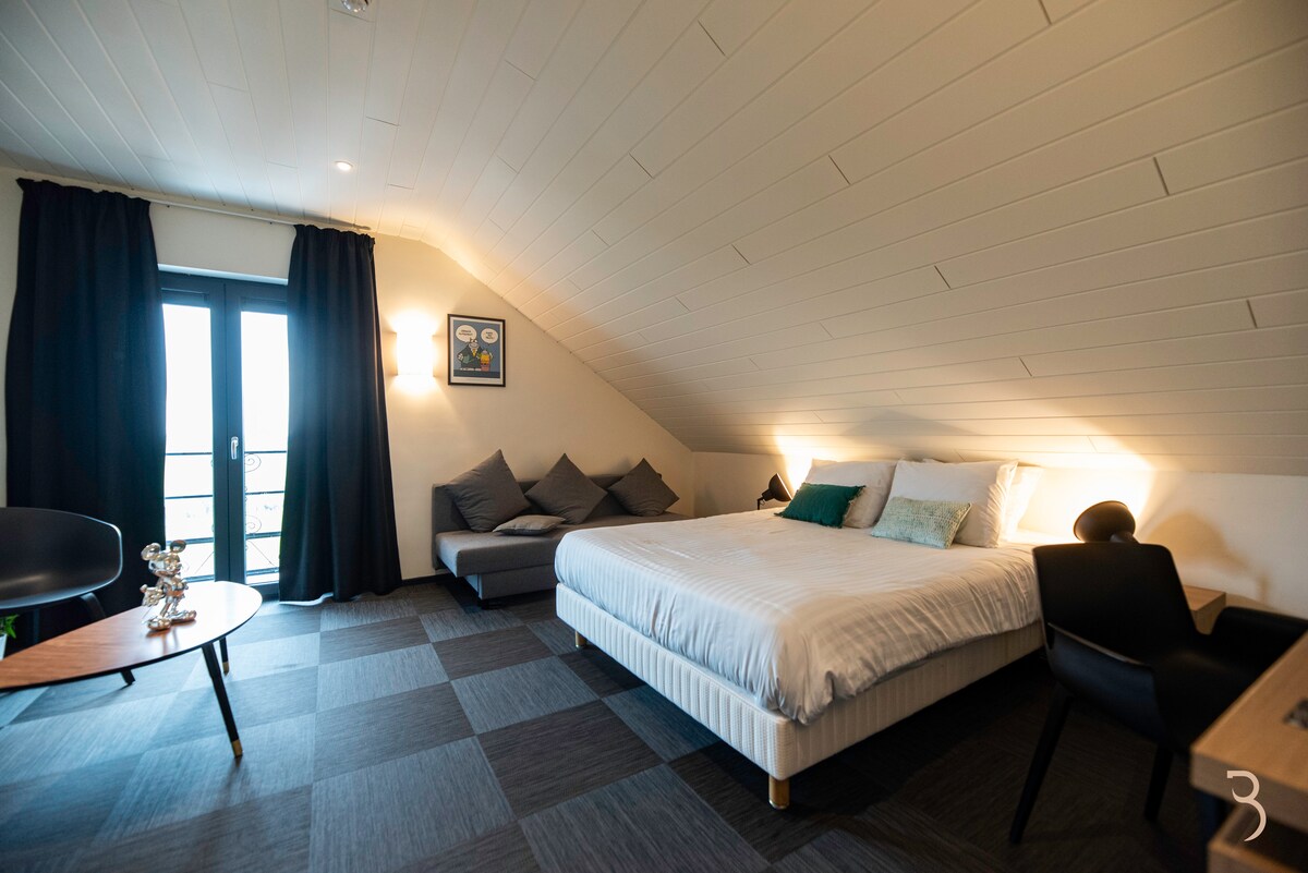 An inviting guest room features a spacious bed dressed in crisp white linens. Comfortable seating is provided with large cushions along a sloped wall. A small table and chair are positioned near the window, where natural light enters through balcony doors covered with dark curtains.