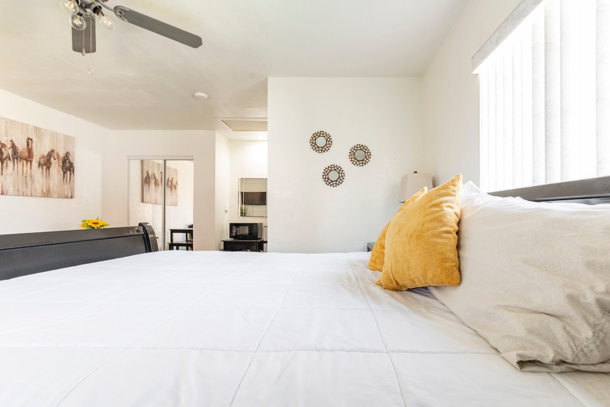 A cozy sleeping area features a spacious bed with white linens and decorative pillows in soft tones. A small kitchenette is visible in the background, complemented by a dining table and chairs. Natural light enters through the window, illuminating the room.